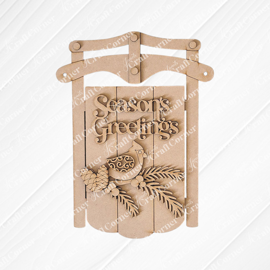 The Season's Greetings Sled Door Hanger from Janet's Craft Corner features carved "Season's Greetings," with a bird, pinecones, and branches on wood. It's an inviting DIY winter decor addition.