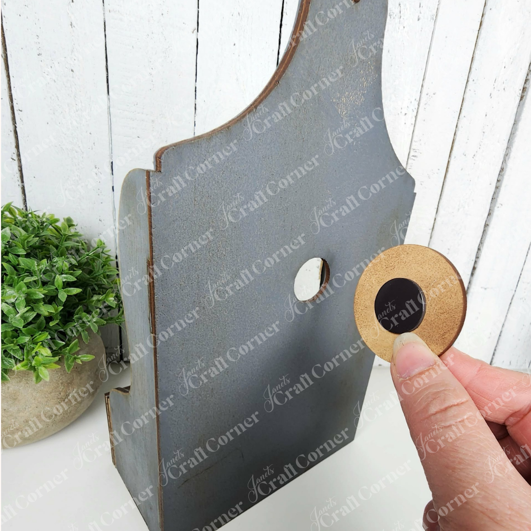 A hand holds a small round magnet near a gray wooden piece with a circular hole from Janet's Craft Corner's Shaker Tea Light Box Interchangeable Set DIY craft kit. A green potted plant accents the farmhouse decor on a white wooden surface.