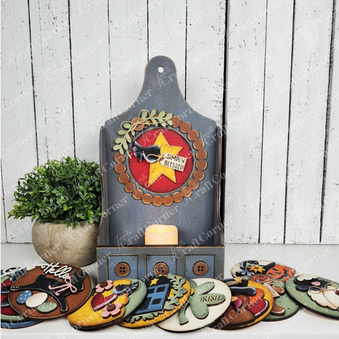 Create a cozy vibe with Janet's Craft Corner Shaker Tea Light Box Interchangeable Set—DIY seasonal home decor kit featuring a wood display, "Simply Blessed" sign, faux greenery, candle, potted plant, and colorful accents on a white background.