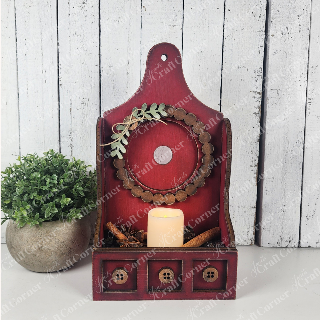 A rustic red wooden shelf with three buttoned drawers displays a beaded wreath, cinnamon sticks, greenery, and Janet's Craft Corner Shaker Tea Light Box Interchangeable Set—seasonal farmhouse decor with shelf sitters and a potted plant.