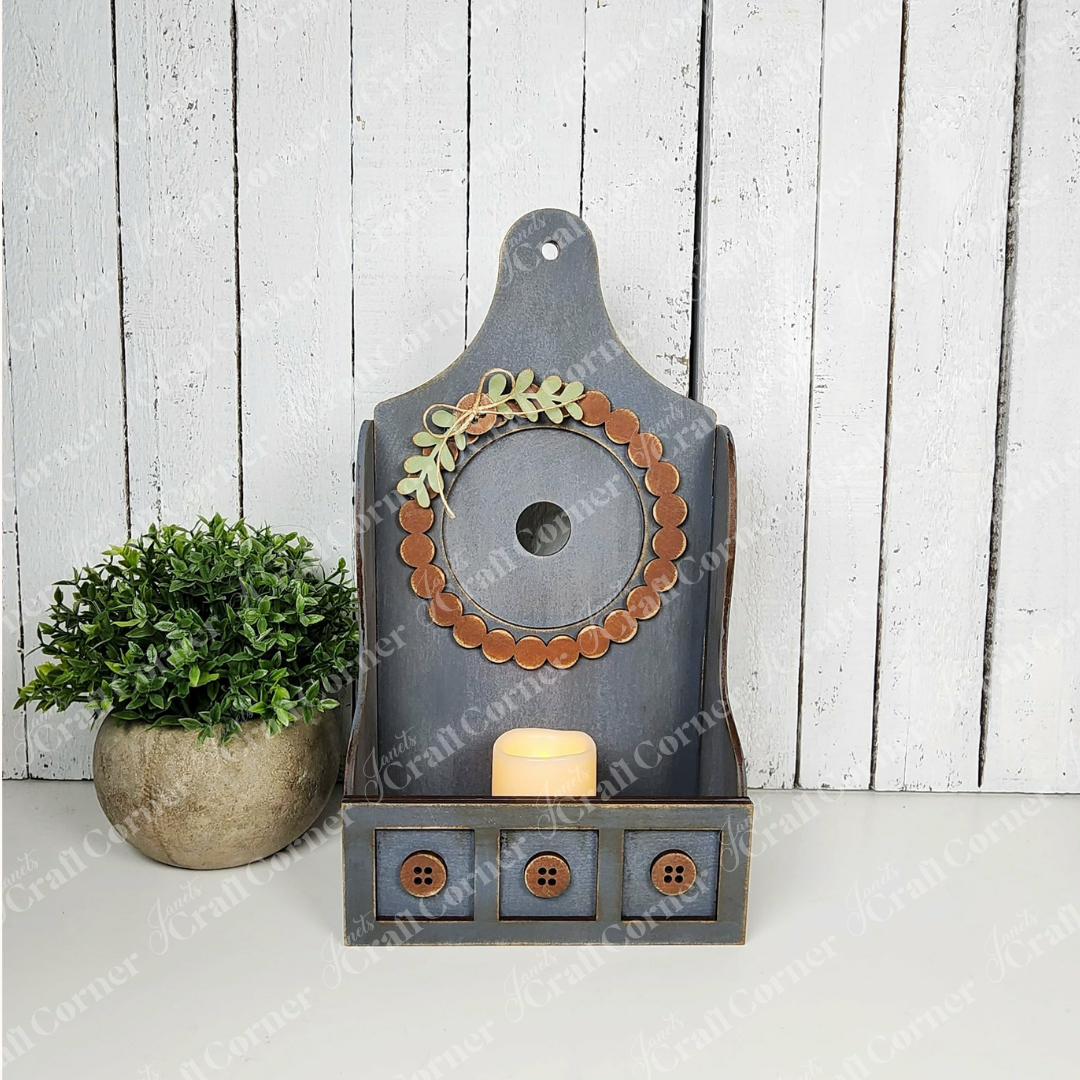 The Shaker Tea Light Box Interchangeable Set by Janet's Craft Corner is a rustic DIY seasonal home decor kit featuring a faux plant, candle, button accents, and wreath detail—ideal for farmhouse style. Set of 1.