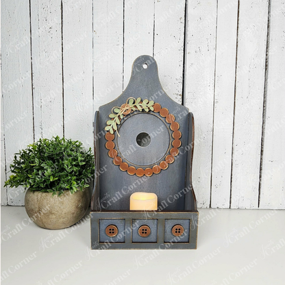 The Shaker Tea Light Box Interchangeable Set by Janet's Craft Corner is a rustic DIY seasonal home decor kit featuring a faux plant, candle, button accents, and wreath detail—ideal for farmhouse style. Set of 1.
