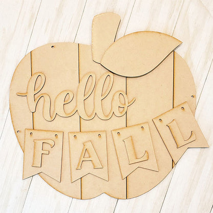 The Shiplap Apple Door Hanger by Janet's Craft Corner is a DIY seasonal home decor craft kit. It features a wooden apple shape with "hello" in script and "FALL" on banner flags, topped with a leaf, all on a light wooden background. Perfect for crafting fun!.