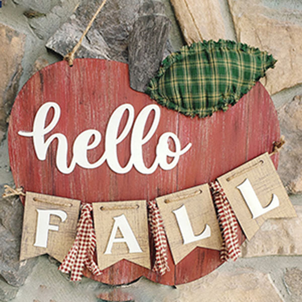 Janet's Craft Corner offers the Shiplap Apple Door Hanger DIY kit, a rustic red apple-shaped sign with white "hello" text and a burlap "FALL" banner, decorated with a green plaid leaf—ideal for hanging against stone.