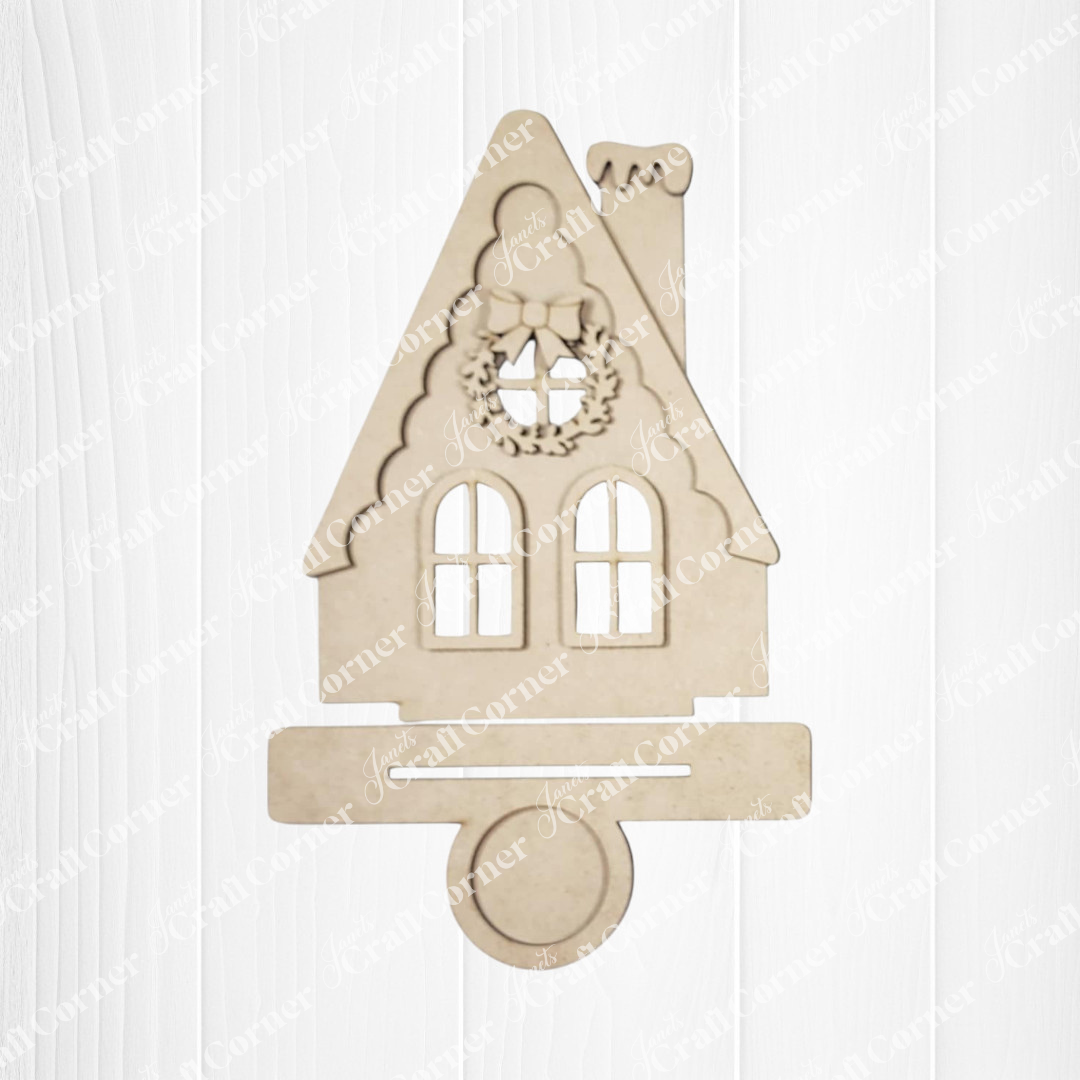 The Victorian Gingerbread House Votive Holder from Janet's Craft Corner is a beige wooden cutout with a steep roof, two windows, chimney, and wreath—a charming DIY seasonal home decor craft kit. Displayed on a white wooden background.