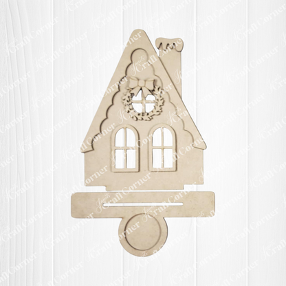 The Victorian Gingerbread House Votive Holder from Janet's Craft Corner is a beige wooden cutout with a steep roof, two windows, chimney, and wreath—a charming DIY seasonal home decor craft kit. Displayed on a white wooden background.