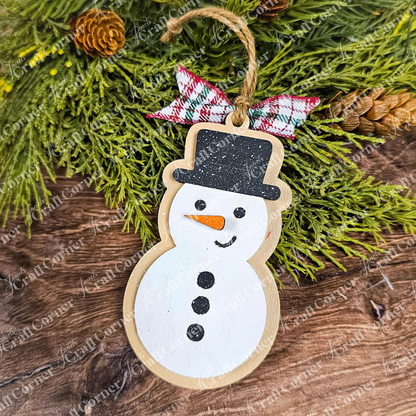A handcrafted gingerbread cookie ornament with a black hat, orange nose, and black buttons hangs from a branch decorated with green foliage and pine cones. A plaid fabric bow adds charm to the Sugar Cookie Ornaments DIY Decoration Kit creation from Janet's Craft Corner, contrasting beautifully against the wooden background.