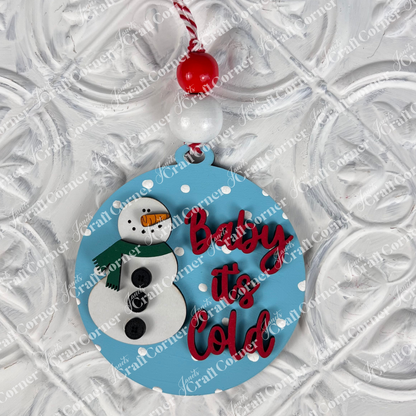 A round Christmas ornament from Janet's Craft Corner's DIY Decoration Kits, featuring a blue design with a snowman and red text stating "Baby it's Cold," hangs from a festive red and white string. This charming decoration, perfect for holiday home décor, is adorned with white polka dots against a textured white background.
