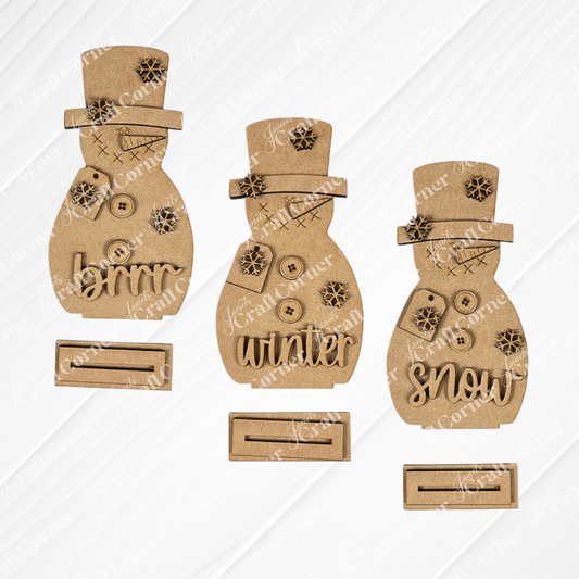 Janet's Craft Corner Snowman Shelf Sitters—three unfinished cardboard snowmen with buttons and "snow," "brrr," and "winter" accessories—are displayed on a white textured surface with stands, perfect for winter décor.