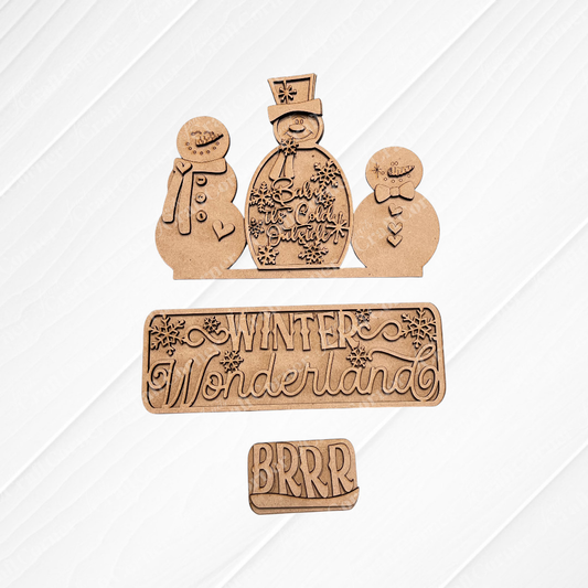 Wooden cutouts on a textured, white square background showcase two snowmen—one adorned with a top hat and scarf, the other smaller. The text "Winter Wonderland" and "BRRR" is displayed in decorative lettering. Ideal for personalized home décor, the burlap surface beneath adds a cozy touch. This is the Winter Wonderland Interchangeable Insert by Janet's Craft Corner.