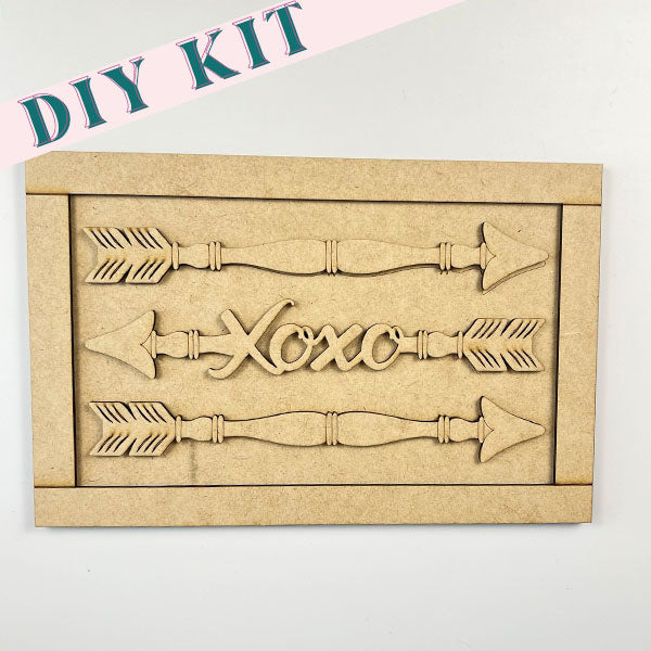 The Spindle Arrows Shelf Leaner from Janet's Craft Corner is an ideal choice for home décor enthusiasts, showcasing two arrows and the word "Xoxo" horizontally arranged in the center, all beautifully framed within a rectangular border.