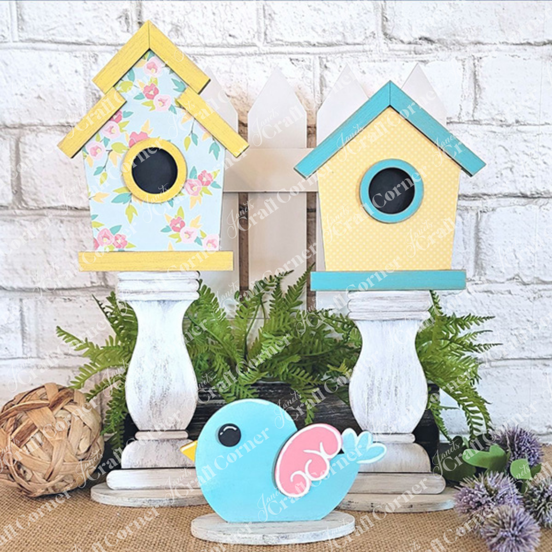 The Spindle Birdhouses Shelf Sitter by Janet's Craft Corner showcases charming floral and polka dot designs atop elegant white stands. Accompanying the setup is an adorable blue wooden bird adorned with a pink swirl. At the base, green ferns and a woven ball are artfully arranged against a white brick backdrop, making it an ideal DIY kit or decorative shelf piece.
