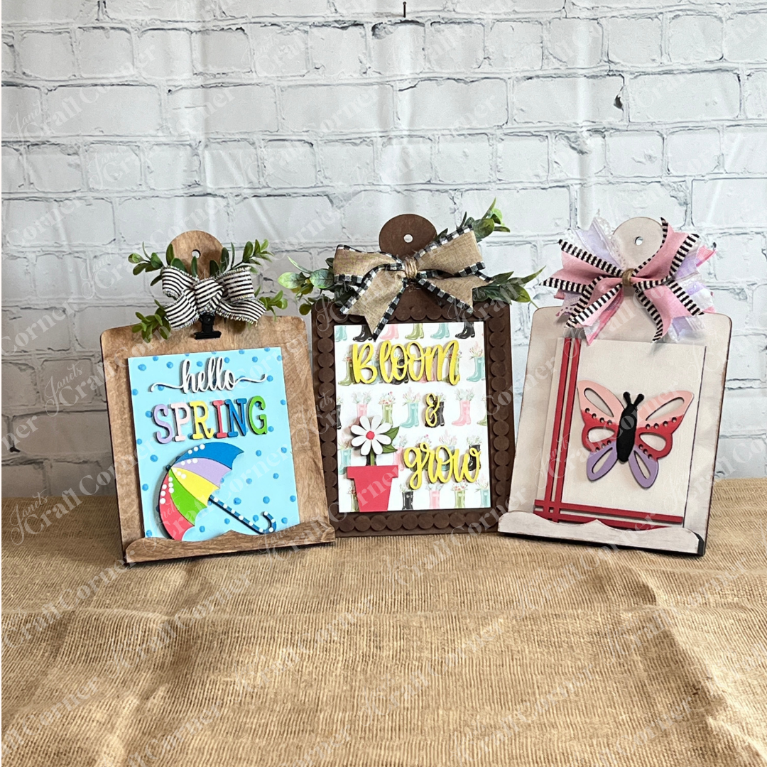 Three mini clipboards from Janet's Craft Corner's Spring Switchable Farmhouse Signs DIY Wood Decor Kit stand on burlap, showcasing cheerful seasonal signs with bows, greenery, and flowers—ideal for tiered tray styling or your spring farmhouse DIY décor.