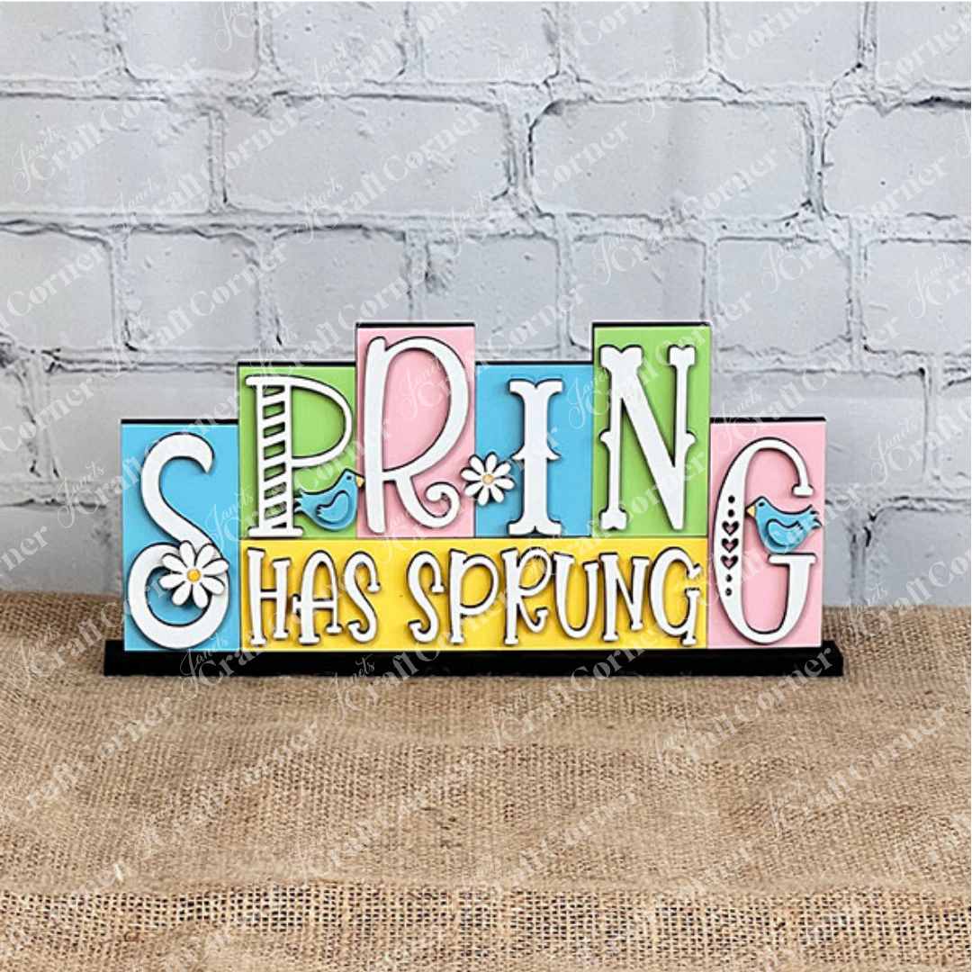 The Hello Spring Mini Word Block from Janet's Craft Corner features vibrant wooden lettering, with playful font variations. Ideal for home décor, it showcases a charming blue bird perched next to the letters "P" and G,  and delightful daisies embellishing the "S" and "I." Positioned against a white background on a burlap surface, it's perfect for adding a touch of spring to any room.