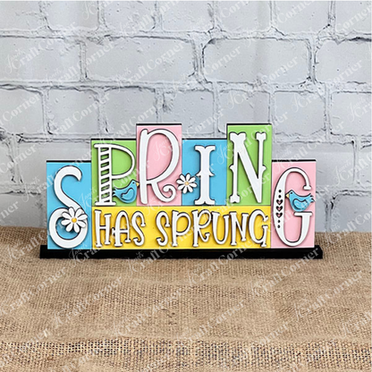 The Hello Spring Mini Word Block from Janet's Craft Corner features vibrant wooden lettering, with playful font variations. Ideal for home décor, it showcases a charming blue bird perched next to the letters "P" and G,  and delightful daisies embellishing the "S" and "I." Positioned against a white background on a burlap surface, it's perfect for adding a touch of spring to any room.