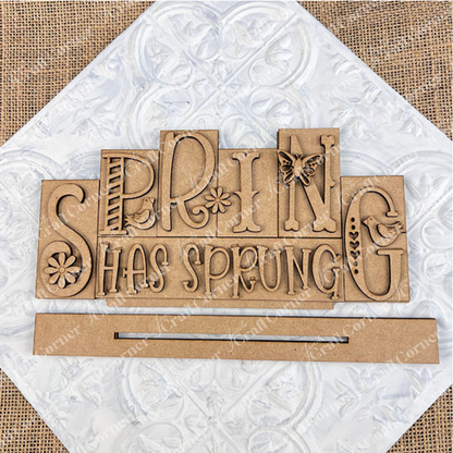 The Hello Spring Mini Word Block from Janet's Craft Corner features vibrant wooden lettering, with playful font variations. Ideal for home décor, it showcases a charming bird perched next to the letters "P" and G, a fun butterfly on the letter "N",  and delightful daisies embellishing the "S" and "I." Positioned against a white background on a burlap surface, it's perfect for adding a touch of spring to any room.