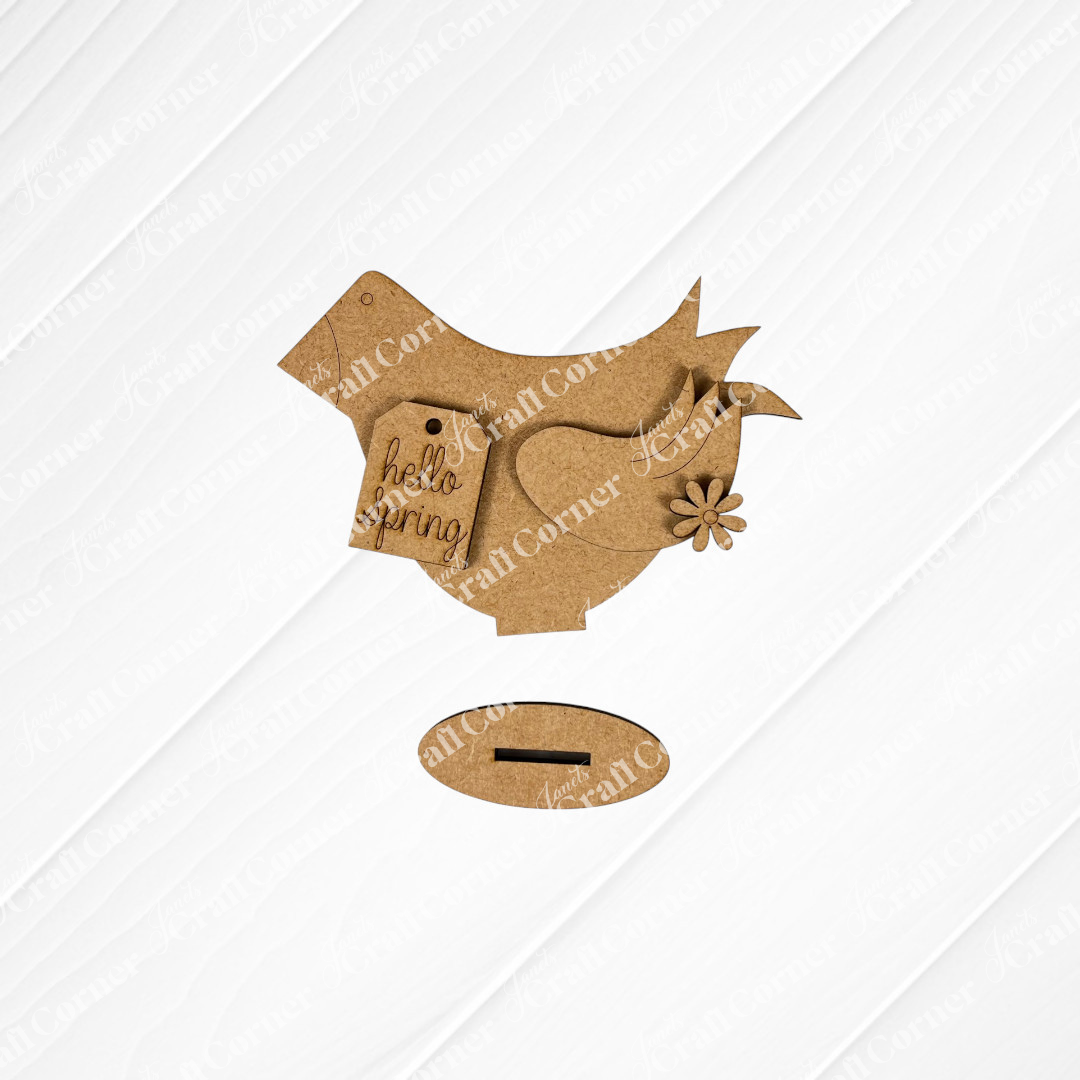 A decorative cardboard bird from Janet's Craft Corner stands on a textured white surface, featuring a charming "hello spring" tag and a small flower. Accompanying it is a small oval piece for a stand, all shown on a white wooden background. This delightful piece perfectly embodies the charm of DIY home décor, making it an ideal addition to your collection of Spring Standing Signs.