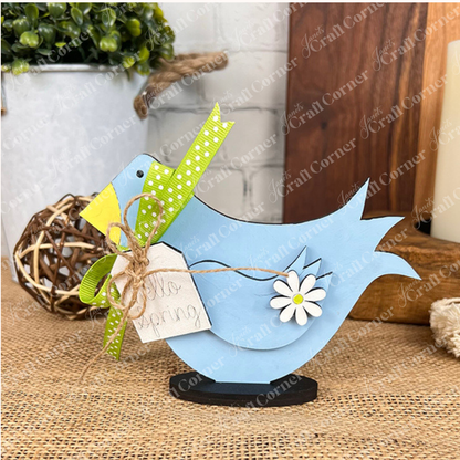 A charming piece from Janet's Craft Corner, the Spring Standing Signs include a blue wooden bird figurine with a yellow beak and green ribbon, prominently displayed on a burlap surface. Featuring delightful flower details and a tag reading "Hello Spring," it adds an inviting touch to your home décor. Surrounding DIY kit elements like potted plants and decorative items complete the scene with grace.