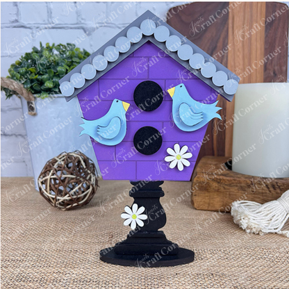 The "Spring Standing Signs" from Janet's Craft Corner is a decorative purple birdhouse ideal for home décor. It showcases two blue birds and white flowers upon a black pedestal, enhanced by the addition of a plant, wooden decor, and candle on a burlap surface to evoke a delightful Spring atmosphere.