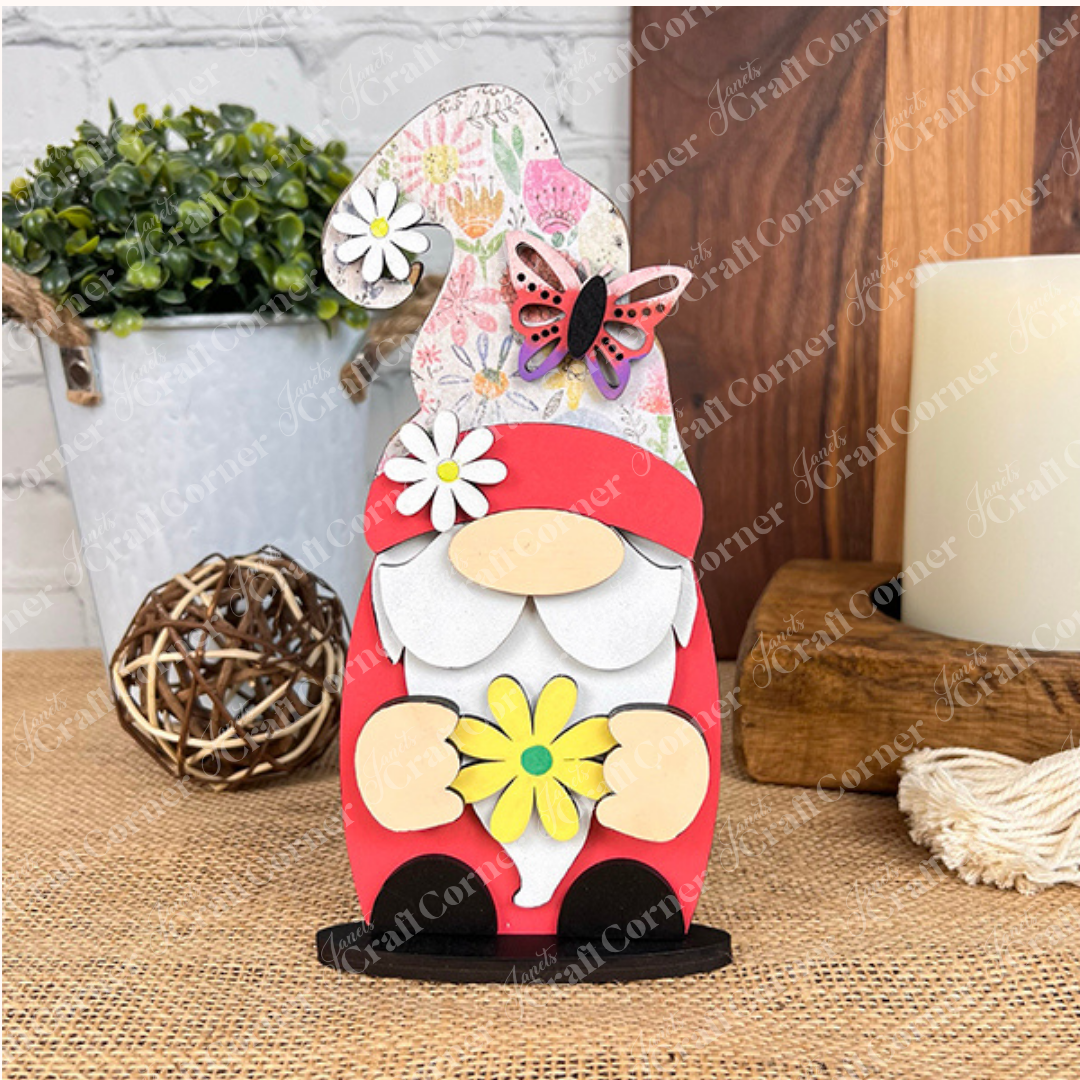 Janet's Craft Corner presents their Spring Standing Signs, featuring a charming decorative wooden gnome adorned with a floral hat and butterfly accent, holding a yellow daisy. It makes an ideal home décor piece, beautifully displayed on a woven surface with a potted plant and candle in the background, all set against a white brick wall.