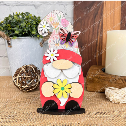 Janet's Craft Corner presents their Spring Standing Signs, featuring a charming decorative wooden gnome adorned with a floral hat and butterfly accent, holding a yellow daisy. It makes an ideal home décor piece, beautifully displayed on a woven surface with a potted plant and candle in the background, all set against a white brick wall.