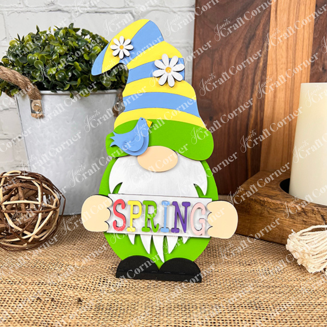 The "Spring Sign Gnome" from Janet's Craft Corner enhances your home décor with its vibrant hues. This colorful wooden figurine features a long white beard and a tall hat adorned with daisies and a blue bird. Holding a "SPRING" sign, it stands amongst plants and decorative items on a woven surface, embodying the spirit of the season.