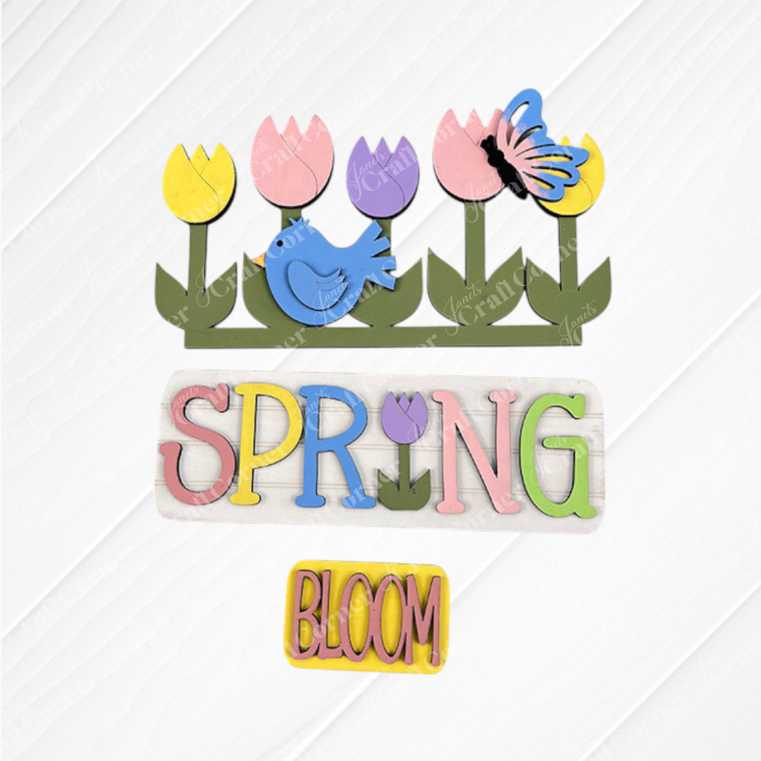 A vibrant wooden art piece from Janet's Craft Corner, designed as an interchangeable insert for home decor, showcases tulips, a blue bird, and a butterfly against a textured background. The words "SPRING" and "BLOOM" are creatively crafted in lively pastel hues. Perfectly suited for adding a splash of seasonal charm to any space!