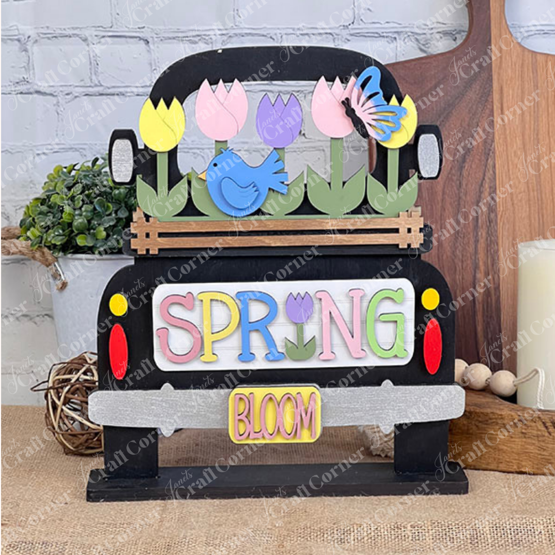 The Spring Tulips Interchangeable Insert from Janet's Craft Corner is a decorative handmade wooden truck adorned with vibrant tulips, a butterfly, and a bird. The word "SPRING" is elegantly displayed on the side with a small "BLOOM" sign beneath it. This charming piece, set against a rustic background, serves as both delightful home decor and an interchangeable insert for various seasons.