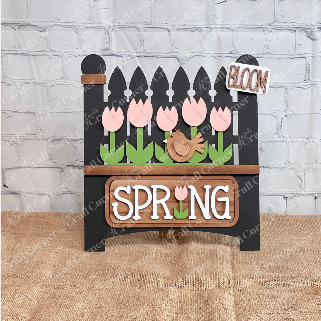 The Spring Tulips Interchangeable Insert from Janet's Craft Corner is a delightful DIY home decor piece. Shaped like the garden fence, it showcases pink tulips and a brown bird alongside the words "Spring Bloom." Set against a rustic background with a woven mat and plants, it's perfect for adding seasonal flair to your space.