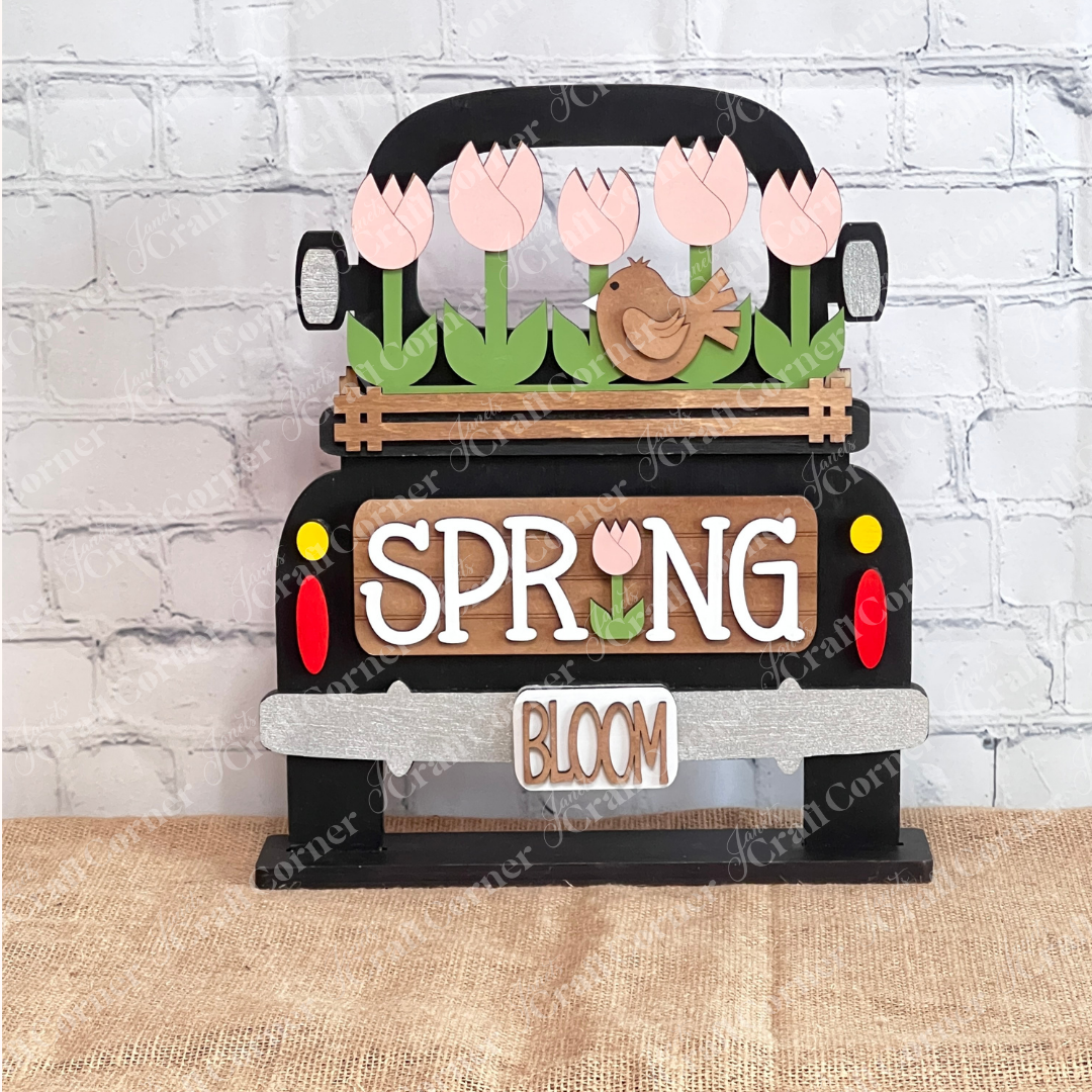 The Spring Tulips Interchangeable Insert from Janet's Craft Corner is a delightful DIY home decor piece. Shaped like the back of a truck, it showcases pink tulips and a brown bird alongside the words "Spring Bloom." Set against a rustic background with a woven mat and plants, it's perfect for adding seasonal flair to your space.