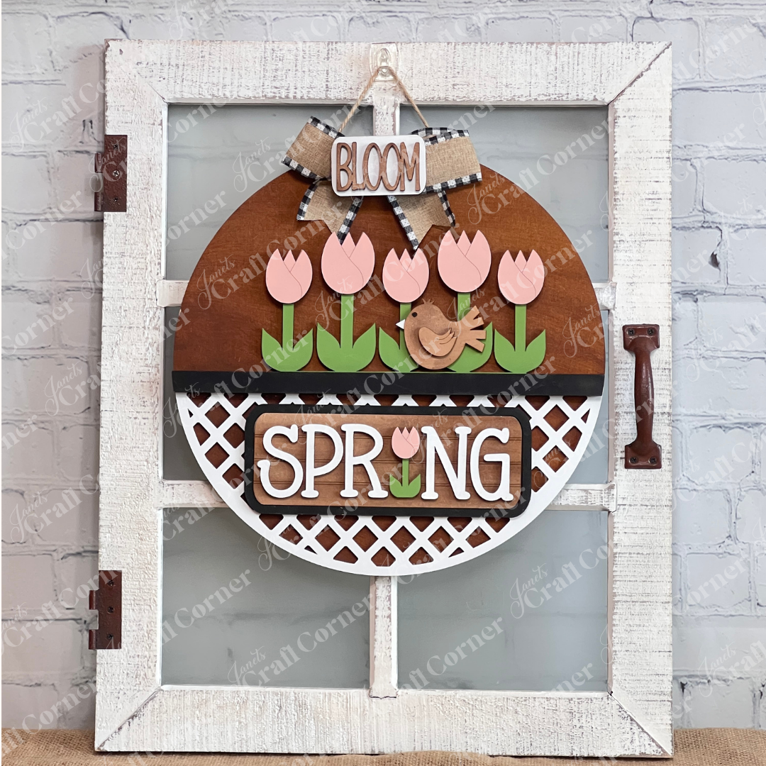 The Spring Tulips Interchangeable Insert from Janet's Craft Corner is a delightful DIY home decor piece. Shaped like the round door hanger on a decorative glass windowed door, it showcases pink tulips and a brown bird alongside the words "Spring Bloom." Set against a rustic background with a woven mat and plants, it's perfect for adding seasonal flair to your space.