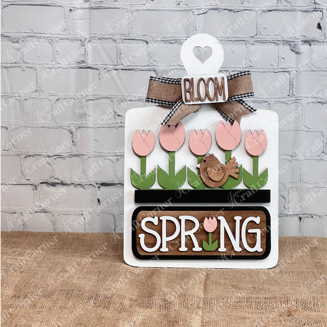 The Spring Tulips Interchangeable Insert from Janet's Craft Corner is a delightful DIY home decor piece. Shaped like the white breadboard, it showcases pink tulips and a brown bird alongside the words "Spring Bloom." Set against a rustic background with a woven mat and plants, it's perfect for adding seasonal flair to your space.