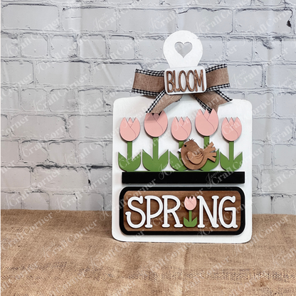 The Spring Tulips Interchangeable Insert from Janet's Craft Corner is a delightful DIY home decor piece. Shaped like the white breadboard, it showcases pink tulips and a brown bird alongside the words "Spring Bloom." Set against a rustic background with a woven mat and plants, it's perfect for adding seasonal flair to your space.