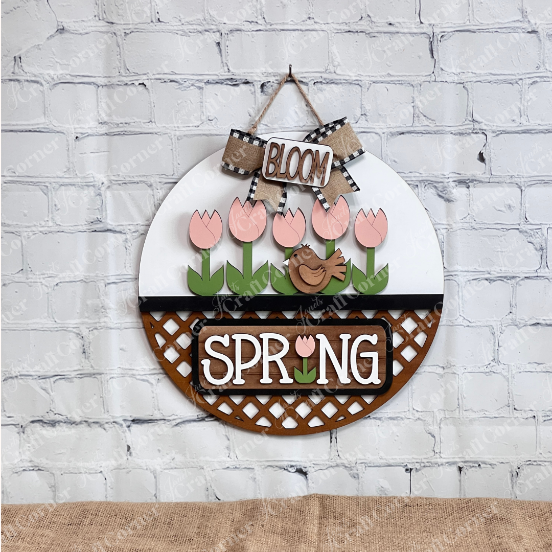 The Spring Tulips Interchangeable Insert from Janet's Craft Corner is a delightful DIY home decor piece. Shaped like the round door hanger, it showcases pink tulips and a brown bird alongside the words "Spring Bloom." Set against a rustic background with a woven mat and plants, it's perfect for adding seasonal flair to your space.