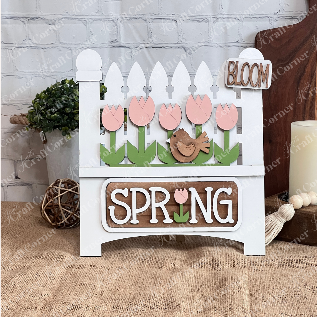 The Spring Tulips Interchangeable Insert from Janet's Craft Corner is a delightful DIY home decor piece. Shaped like the picket fence, it showcases pink tulips and a brown bird alongside the words "Spring Bloom." Set against a rustic background with a woven mat and plants, it's perfect for adding seasonal flair to your space.