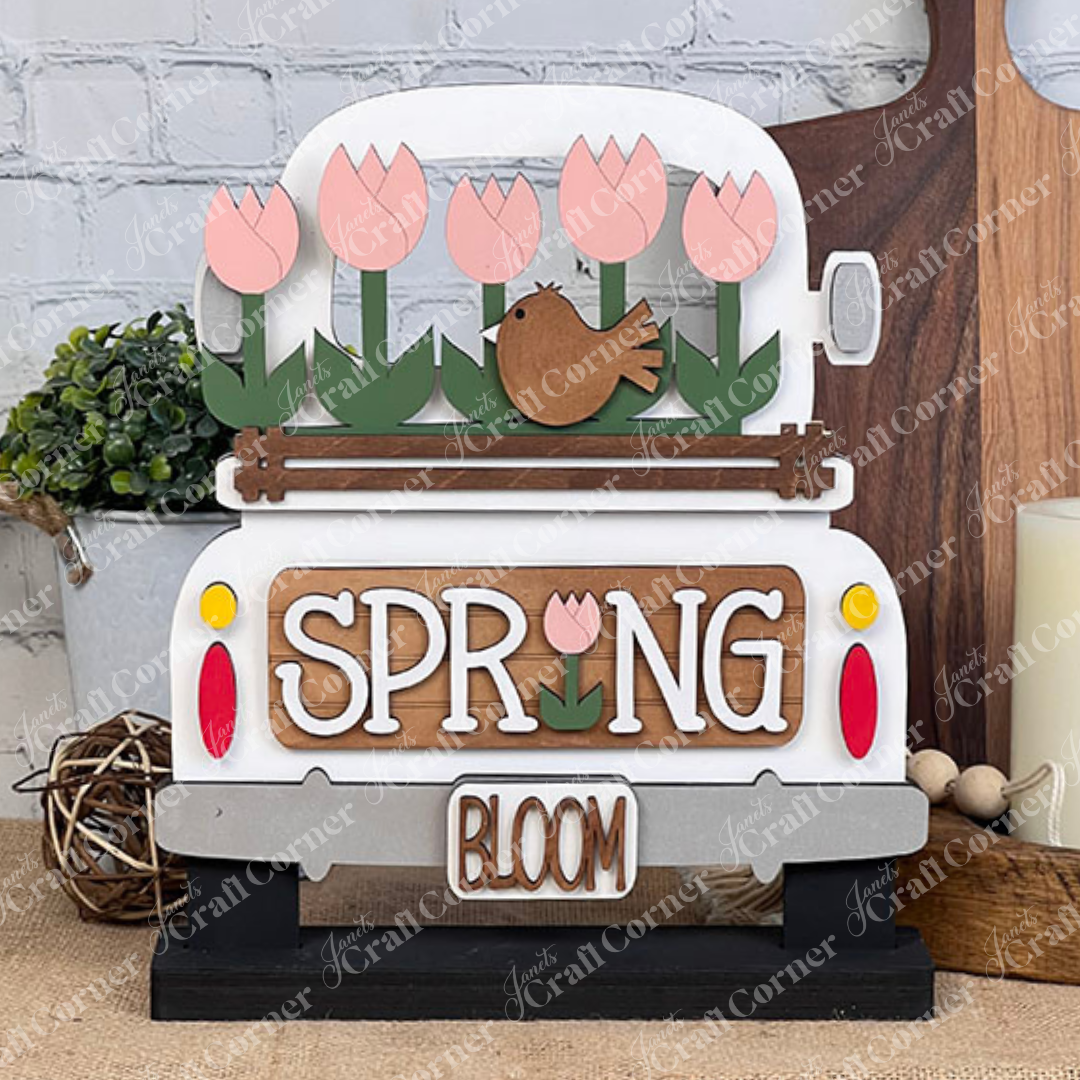 The Spring Tulips Interchangeable Insert from Janet's Craft Corner is a delightful DIY home decor piece. Shaped like the back of a truck, it showcases pink tulips and a brown bird alongside the words "Spring Bloom." Set against a rustic background with a woven mat and plants, it's perfect for adding seasonal flair to your space.