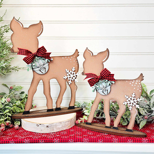 Two standing deer from Janet's Craft Corner, part of the Standing Deer Shelf Sitter DIY Home Decor Kit - Set of 2, proudly stand on a white surface. These wooden decorations boast red plaid bows and snowflake ornaments and are beautifully complemented by green foliage and a red-checkered cloth adorned with white snowflakes.