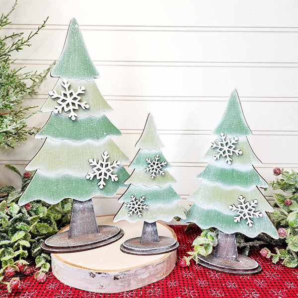 A set of three standing trees from Janet's Craft Corner, part of their DIY Home Decor Craft Kit collection, features layered green segments adorned with delicate white snowflakes. These decorative wooden trees vary in height, offering a charming hand-painted appearance as they grace a red plaid surface amidst surrounding greenery, all against a backdrop of light-colored paneling.