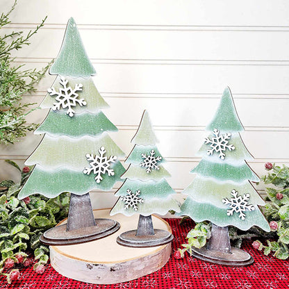 A set of three standing trees from Janet's Craft Corner, part of their DIY Home Decor Craft Kit collection, features layered green segments adorned with delicate white snowflakes. These decorative wooden trees vary in height, offering a charming hand-painted appearance as they grace a red plaid surface amidst surrounding greenery, all against a backdrop of light-colored paneling.