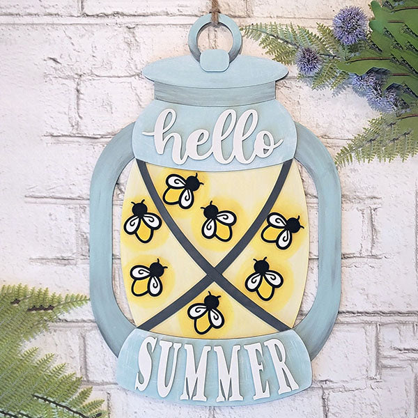 Add a touch of charming farmhouse style to your home decor with the Summer Lantern Door Hanger from Janet's Craft Corner. This decorative wooden sign, shaped like a lantern and featuring the words "hello summer," showcases cute bees and a cheerful yellow background. Perfect for gracing a white brick wall, it's beautifully complemented by vibrant greenery.