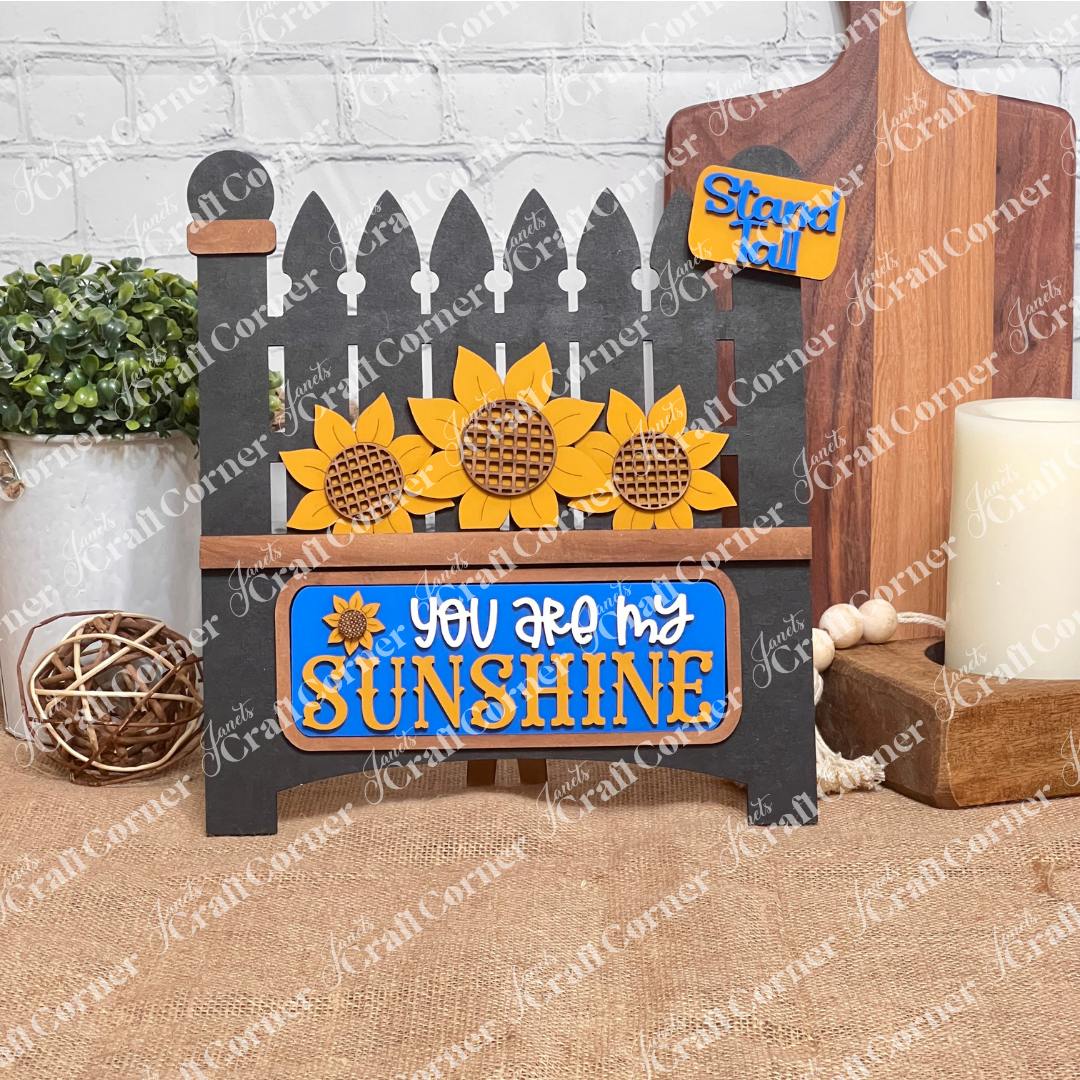 The Sunflower Interchangeable Insert from Janet's Craft Corner is a DIY home decor kit featuring a black garden gate cutout adorned with sunflowers and the phrase "You Are My Sunshine" in yellow and white, plus a small "Stand Tall" tag. Ideal for fans of interchangeable decorations, it complements any setting with plants, candles, or cutting boards.