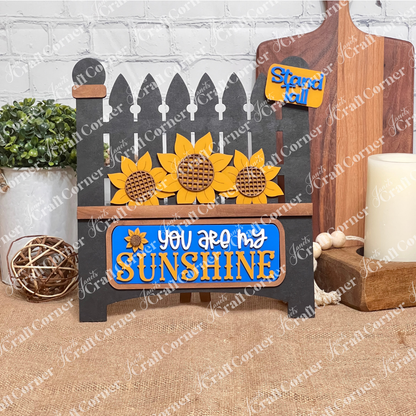 The Sunflower Interchangeable Insert from Janet's Craft Corner is a DIY home decor kit featuring a black garden gate cutout adorned with sunflowers and the phrase "You Are My Sunshine" in yellow and white, plus a small "Stand Tall" tag. Ideal for fans of interchangeable decorations, it complements any setting with plants, candles, or cutting boards.