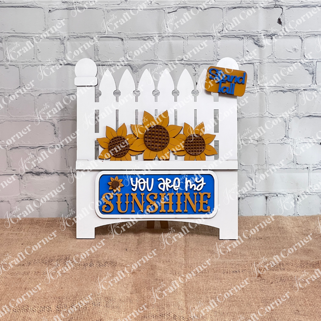 The Sunflower Interchangeable Insert from Janet's Craft Corner is a DIY home decor kit featuring a white picket fence cutout adorned with sunflowers and the phrase "You Are My Sunshine" in yellow and white, plus a small "Stand Tall" tag. Ideal for fans of interchangeable decorations, it complements any setting with plants, candles, or cutting boards.