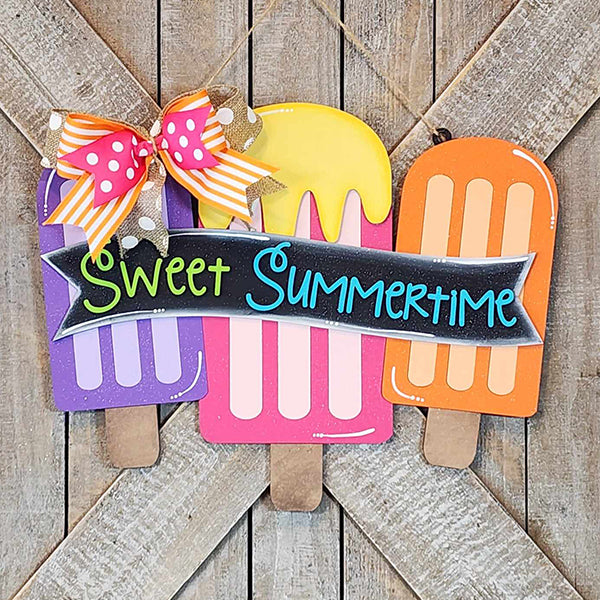 The Sweet Summertime Door Hanger by Janet's Craft Corner showcases three vibrant ice cream popsicles in shades of purple, pink, and orange, accentuated with lively lettering. It features a charming red and orange polka dot bow on the top left corner against a textured wooden background, making it perfect for home décor or a delightful DIY kit project.