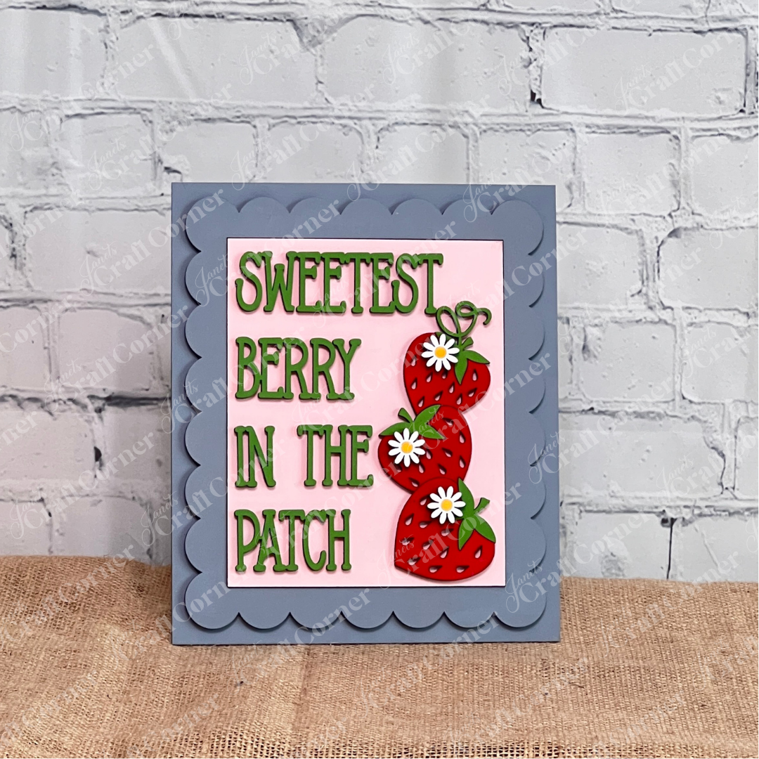 Janet's Craft Corner Single Scallop Trim Farmhouse Frame features "Sweetest Berry In The Patch" in green, with two strawberries and white daisies on a pink background, propped against a white brick wall.