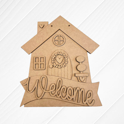The Janet's Craft Corner Welcome House Wall Hanger is a wooden DIY home decor kit featuring a house cutout with two windows, a round-topped door with a heart wreath, and “Welcome”—ideal for brightening any entryway. Set of 1.