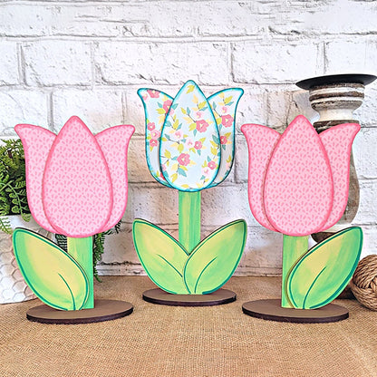 A trio of Janet's Craft Corner Tulip Trio Shelf Sitters is displayed elegantly on a table with a brick wall backdrop. Each tulip showcases distinct patterns and vibrant colors, complemented by green leaves on the stems, crafting a lively and artistic home décor feature.