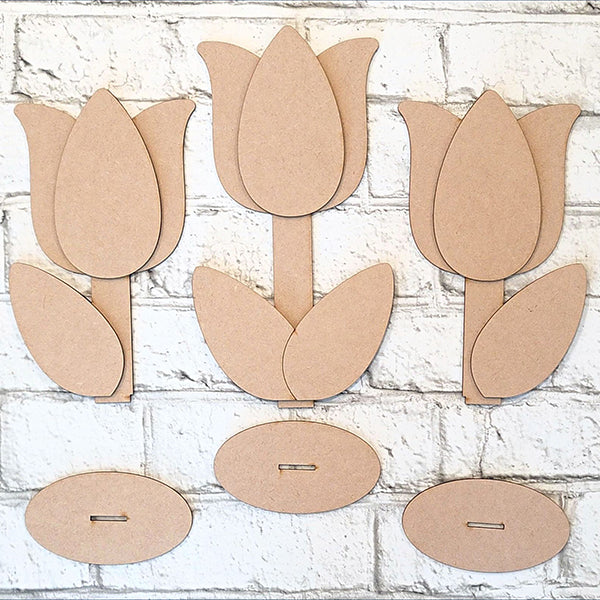 Against a white brick background, the Tulip Trio Shelf Sitters by Janet's Craft Corner are displayed. This DIY kit features three cardboard tulip cutouts with separate petals, two leaves each, stems, and oval bases for easy home décor assembly.