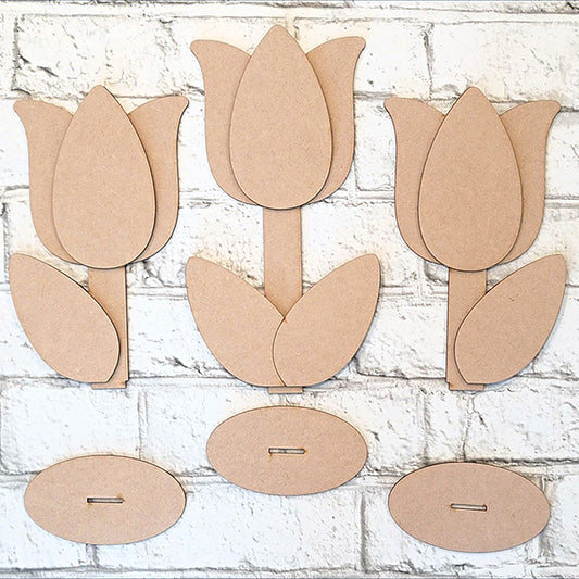 Against a white brick background, the Tulip Trio Shelf Sitters by Janet's Craft Corner are displayed. This DIY kit features three cardboard tulip cutouts with separate petals, two leaves each, stems, and oval bases for easy home décor assembly.