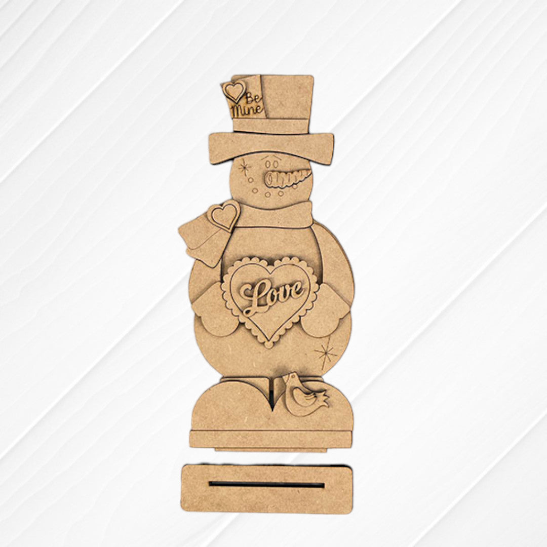 The Valentine Snowman Shelf Sitter by Janet's Craft Corner is a DIY decor craft kit that includes a wooden snowman with heart-shaped "Love" and "Be Mine" details, on a rectangular base with a small bird figure against a textured white and burlap background. Perfect for Valentine's Day decorating.